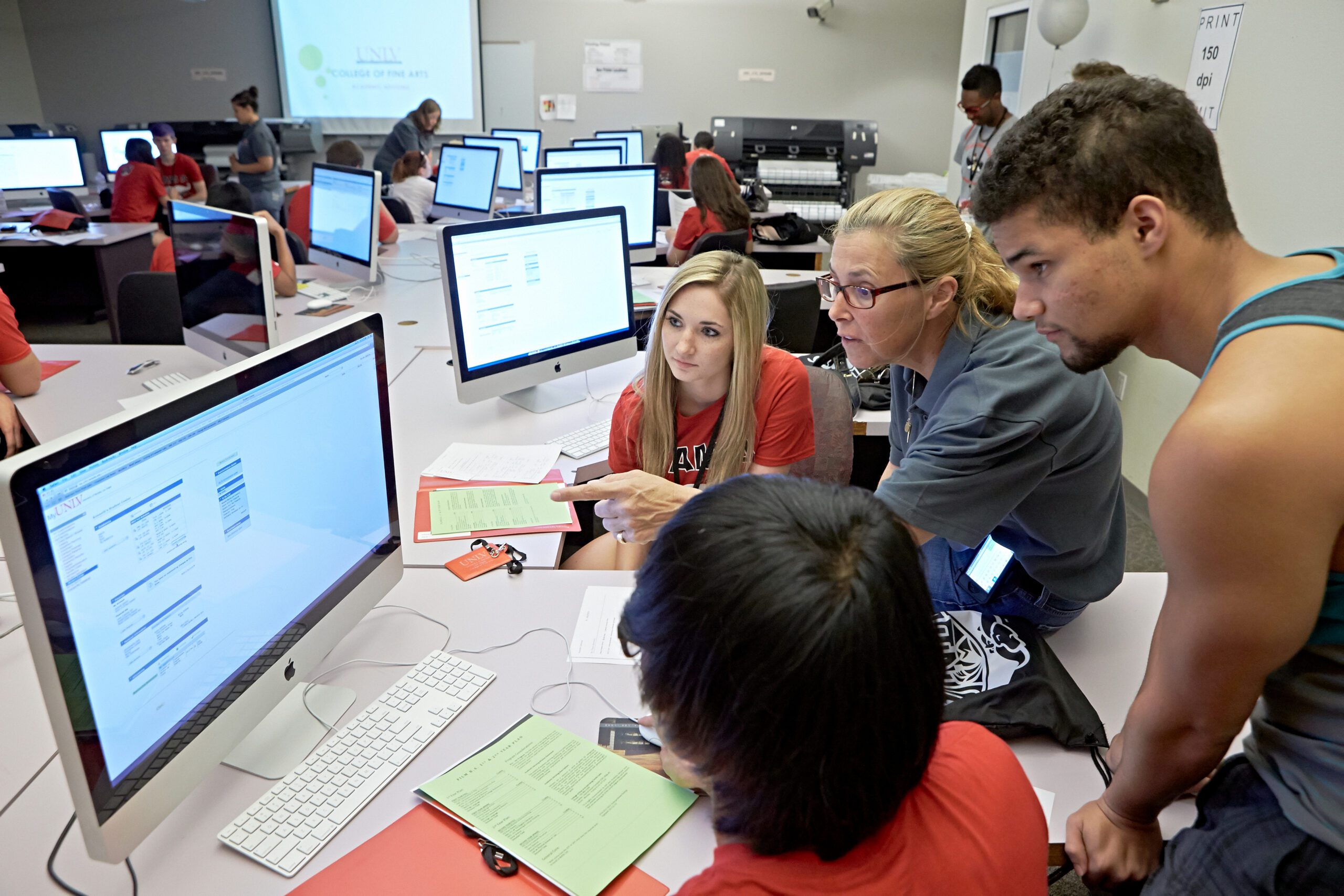 An instructor and three students huddle around a computer monitor. The instructor points to the screen.