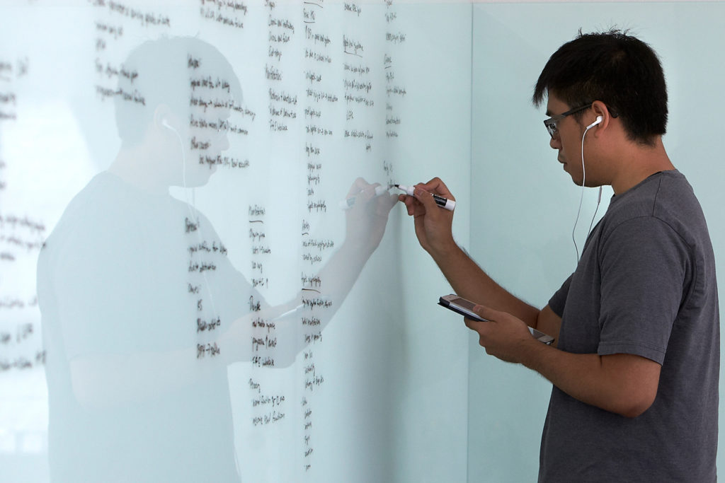 A student adds a line of text on a large whiteboard. Most of the space on the wall-length whitebard is covered with writing.