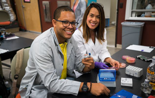 A pair of lab workers smile for the camera. The worker in the foreground holds up a pair of vials in one hand.