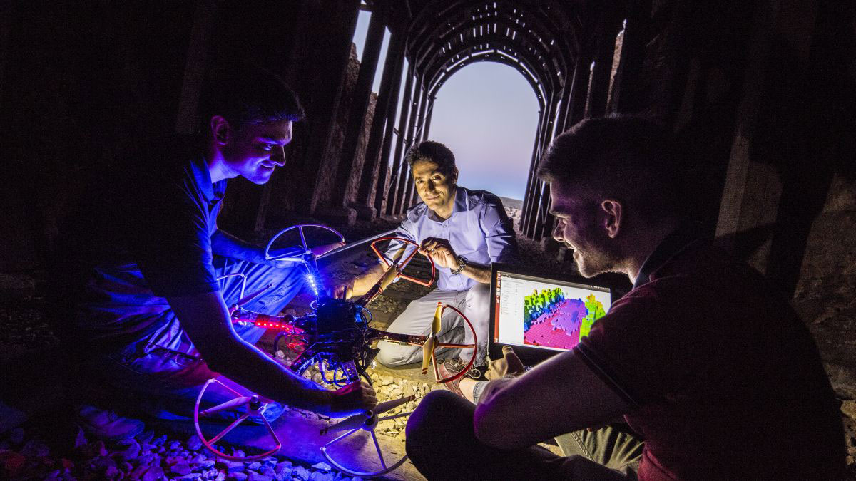 A trio of technicians huddle in a tunnel around a laptop and a connected quadcopter drone.